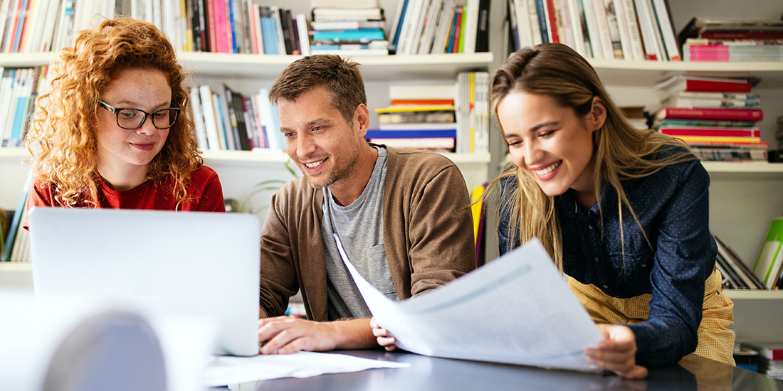 Das Bild zeigt zwei Studentinnen und einen Studenten, die gemeinsam in einer Bibliothek am Tisch sitzen und suchend in eine Zeitung und auf einen Laptop schauen.